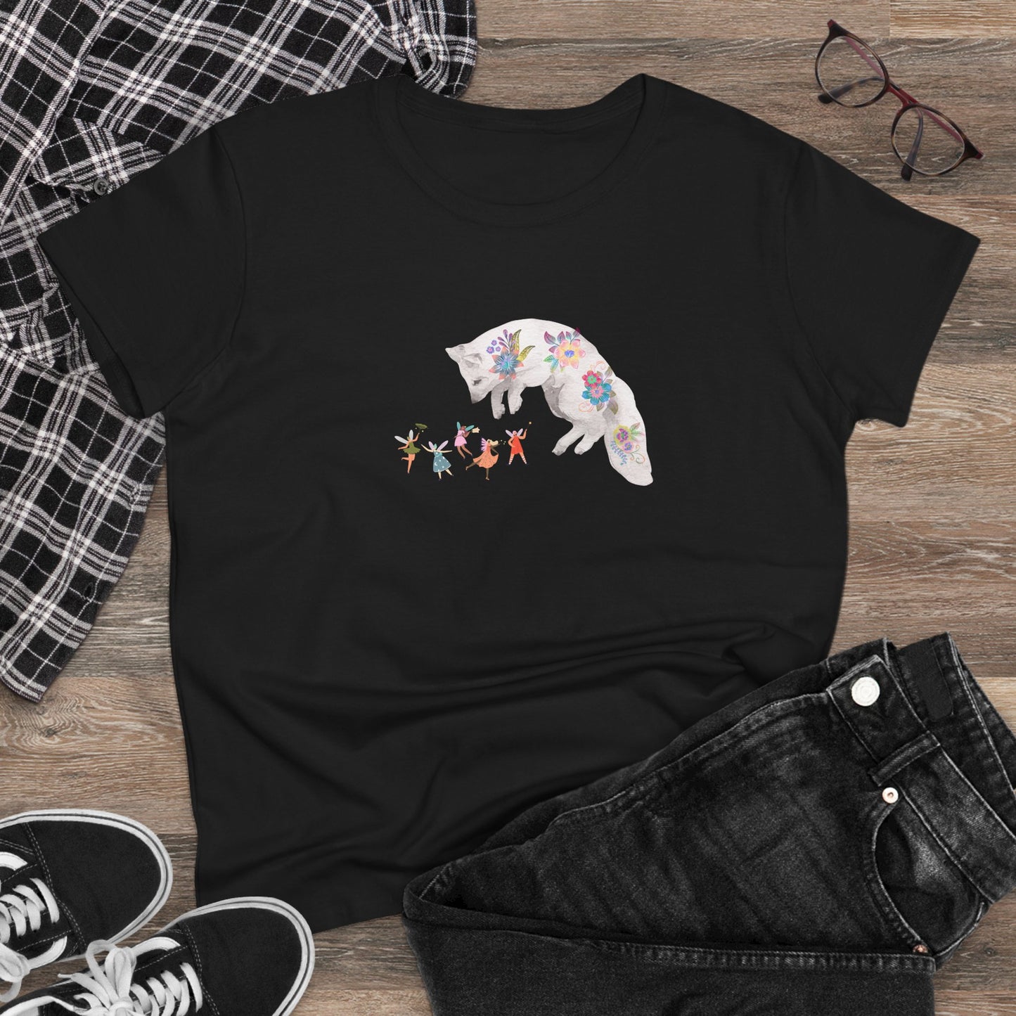 Black t-shirt with a colorful white fox and dancing winged fairies graphic on a wooden surface with jeans and sneakers.