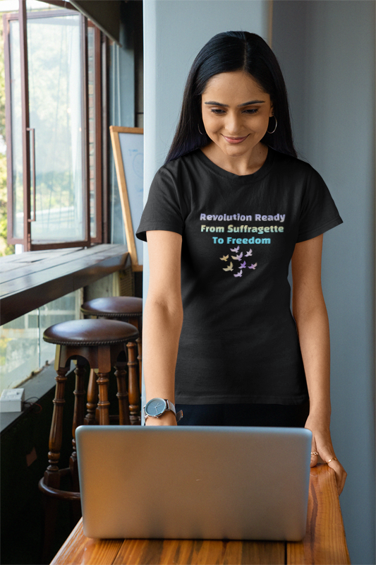 Woman wearing a black t-shirt with a colorful design and text, standing in a room with a window and wooden furniture.