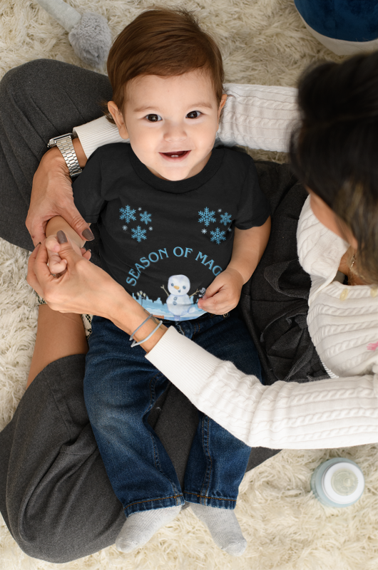 Baby wearing a black shirt with blue snowflakes and text, sitting on a person's lap on a carpeted floor.
