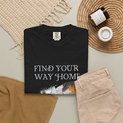 A folded black T-shirt with the phrase 'FIND YOUR WAY HOME' in white serif letters and a small scenic graphic near the bottom, styled in a flat lay with beige pants, a woven blanket, a candle jar, and a small cup on a light neutral background.