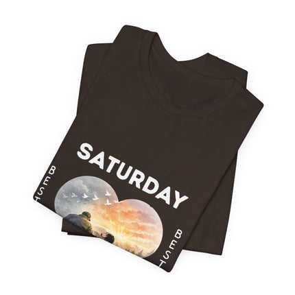 A dark brown folded t-shirt with a graphic print featuring the word 'SATURDAY' in white, alongside a circular landscape illustration of a sunrise and birds, on a plain white background.
