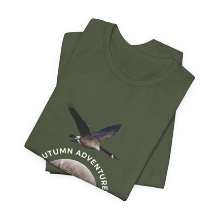 An olive green folded t-shirt featuring a graphic of a flying Canada goose and the text 'AUTUMN ADVENTURE' in white, set against a plain white background.