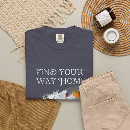 A folded dark slate blue t-shirt with the text 'FIND YOUR WAY HOME' and a winter landscape graphic, styled in a flat lay with beige pants, a knit blanket, a candle, and a woven placemat on a light gray background.