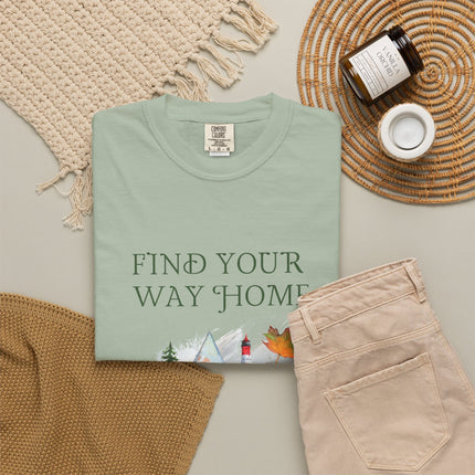 A folded sage green T-shirt with the phrase 'FIND YOUR WAY HOME' in white serif letters and a small scenic graphic near the bottom, styled in a flat lay with beige pants, a woven blanket, a candle jar, and a small cup on a light neutral background.