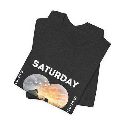 A black folded t-shirt with a graphic print on a white background. The graphic features the word 'SATURDAY' in bold white letters above a heart showing birds and a sunset..