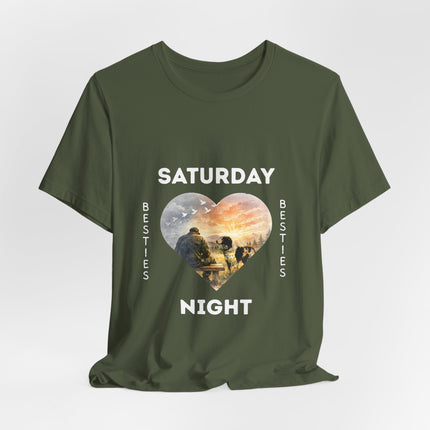 A olive green short-sleeve t-shirt with a graphic print on the front. The design features the text 'SATURDAY' and 'NIGHT' in white capital letters framing a heart-shaped illustration of veteran on a bench with his loving dog staring at him with love.
