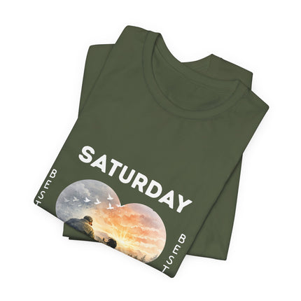 A folded olive green t-shirt with a graphic print on the front. The print features the word 'SATURDAY' in bold white letters above two circular scenic illustrations of a moon and a sunset, with small white birds flying across the sky.