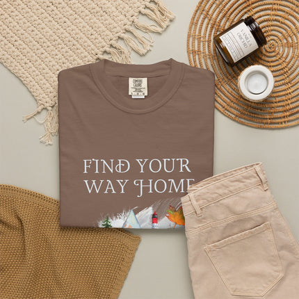 A folded brown T-shirt with the phrase 'FIND YOUR WAY HOME' in white serif letters and a small scenic graphic near the bottom, styled in a flat lay with beige pants, a woven blanket, a candle jar, and a small cup on a light neutral background.