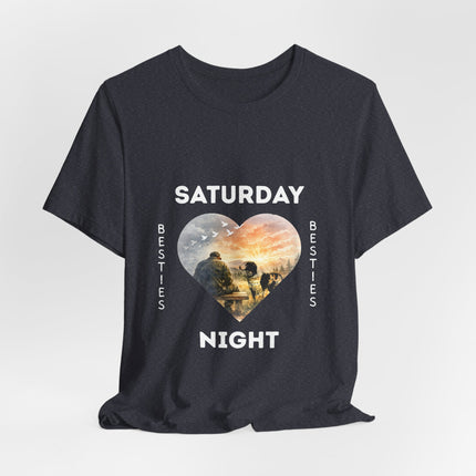 A dark charcoal short-sleeve t-shirt with a graphic print on the front. The design features the text 'SATURDAY' and 'NIGHT' in white capital letters framing a heart-shaped illustration of veteran on a bench with his loving dog staring at him with love.