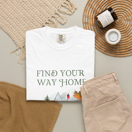 A folded white T-shirt with the phrase 'FIND YOUR WAY HOME' in white serif letters and a small scenic graphic near the bottom, styled in a flat lay with beige pants, a woven blanket, a candle jar, and a small cup on a light neutral background.