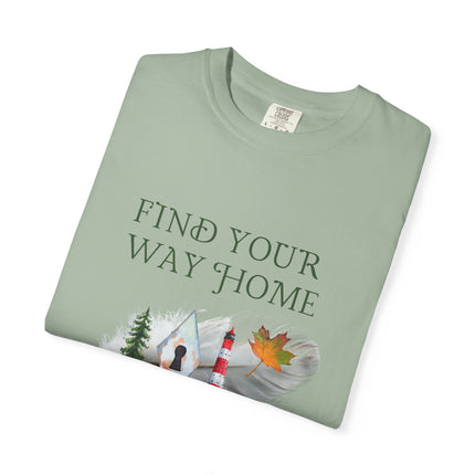 A folded sage green crewneck t-shirt with a graphic print that reads 'FIND YOUR WAY HOME' above an illustration of a lighthouse, a tree, a keyhole, and a maple leaf, shown on a plain white background.