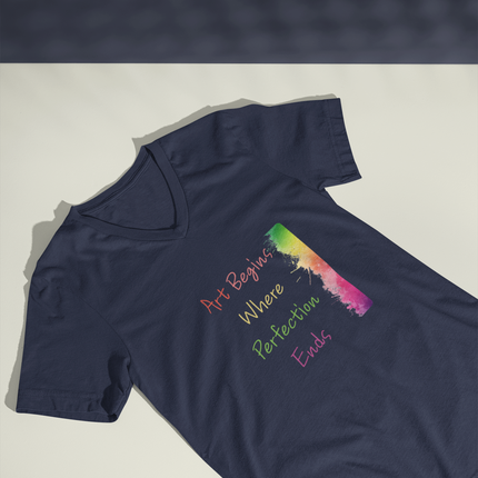 A navy blue V-neck t-shirt laid flat on a light gray surface, featuring a colorful graphic with the text 'Art Begins Where Perfection Ends' and a vertical rainbow-striped brushstroke design.