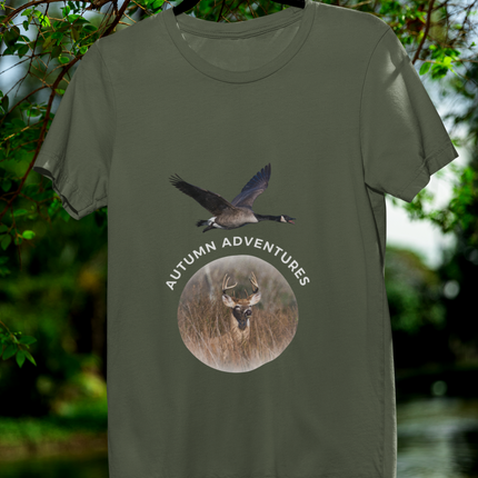 An olive green short-sleeve t-shirt hanging on a black hanger against a blurred outdoor background of green foliage and water. The shirt features a graphic print with a flying goose above a circular deer head and the words 'AUTUMN ADVENTURES'.