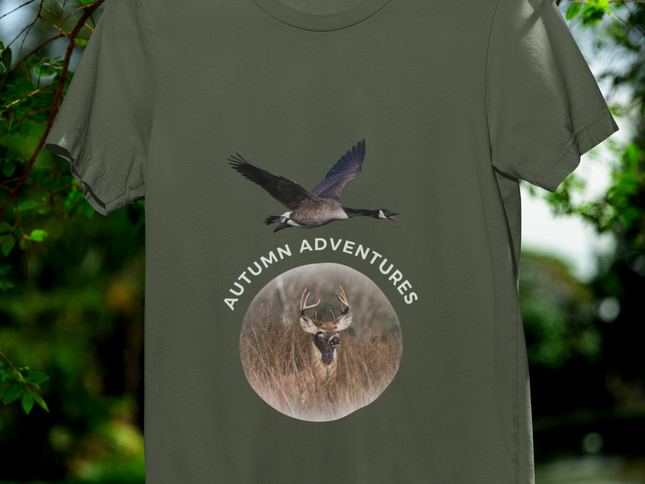 An olive green short-sleeve t-shirt hanging on a black hanger against a blurred outdoor background of green foliage and water. The shirt features a graphic print with a flying goose above a circular deer head and the words 'AUTUMN ADVENTURES'.