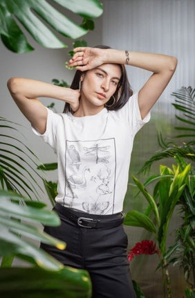A woman posing in a white graphic t-shirt with black line drawings of a dragonfly, swan, and deer, paired with black pants, surrounded by lush green tropical plants.