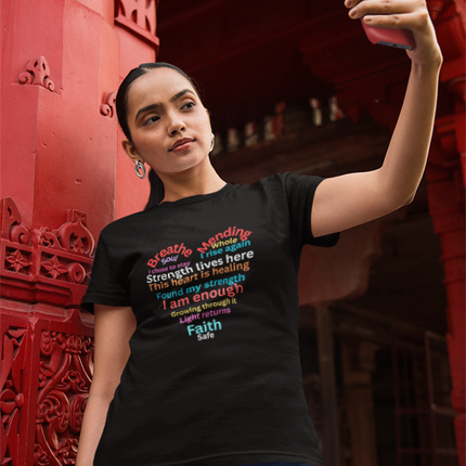 Woman taking a selfie wearing a black t-shirt with colorful text in front of a red architectural background.