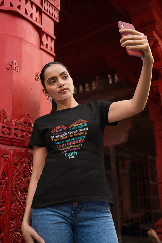 Woman taking a selfie wearing a black t-shirt with colorful text in front of a red architectural background.