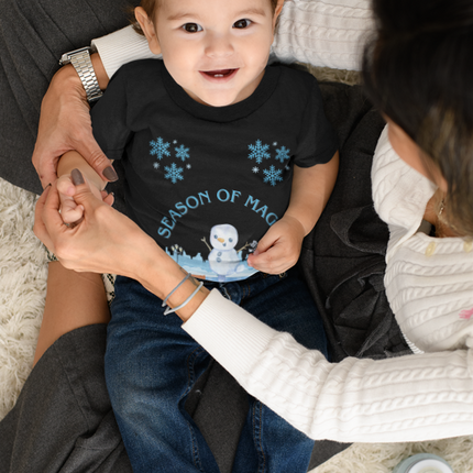 Baby wearing a black shirt with blue snowflakes and text, sitting on a person's lap on a carpeted floor.