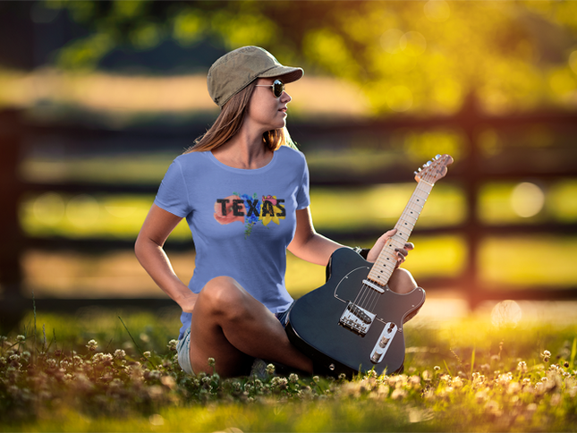 Woman with a guitar sitting on the grass wearing a blue t-shirt that says TEXAS displaying wildflowers behind the print.