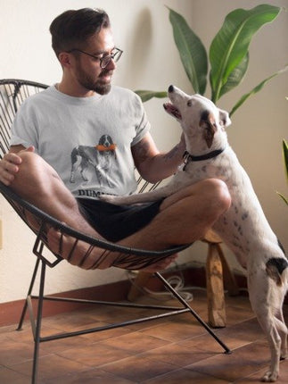 A man wearing a light gray graphic t-shirt with a dog print sits in a black wire chair, petting a white dog with black spots. The setting is indoors with a wooden floor and a large green plant in the background.