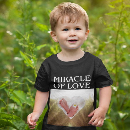 Child wearing a black t-shirt with 'Miracle of Love' design in a natural setting